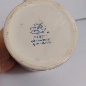 May include: A close-up of the bottom of a white ceramic pitcher with blue lettering that reads "Delfts Handwork Holland". The pitcher has a slightly textured surface and a blue decorative design.