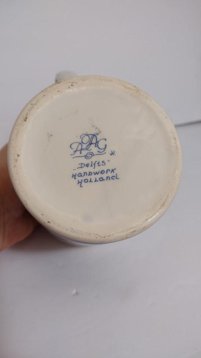 May include: A close-up of the bottom of a white ceramic pitcher with blue lettering that reads "Delfts Handwork Holland". The pitcher has a slightly textured surface and a blue decorative design.
