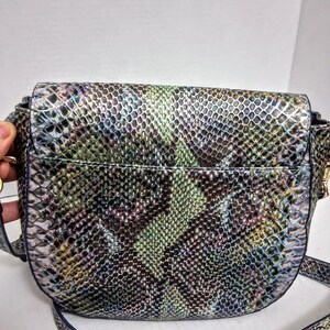 May include: A crossbody bag featuring a textured snakeskin pattern in shades of iridescent green, purple, blue, and black. The bag has a rounded flap closure, a structured silhouette, and a matching adjustable shoulder strap attached to the sides.