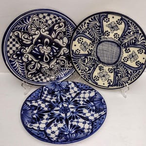 May include: Three ceramic plates with intricate blue and white floral designs. The plates vary in size and pattern, with detailed scrollwork and symmetrical arrangements. The plates are displayed on clear stands, showcasing their decorative nature.