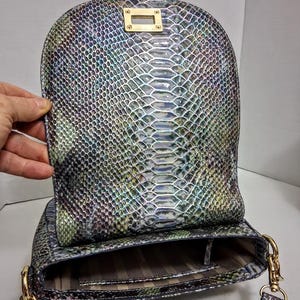 May include: A shoulder bag featuring an iridescent snakeskin pattern in shades of green, blue, and purple. The bag has a rounded flap with a gold-tone rectangular turn-lock closure and gold-tone metal hardware attachments for a shoulder strap.