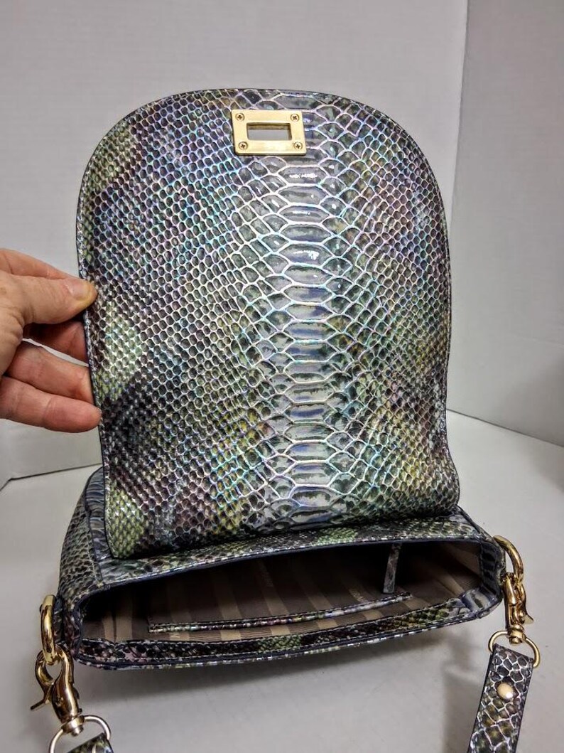 May include: A shoulder bag featuring an iridescent snakeskin pattern in shades of green, blue, and purple. The bag has a rounded flap with a gold-tone rectangular turn-lock closure and gold-tone metal hardware attachments for a shoulder strap.