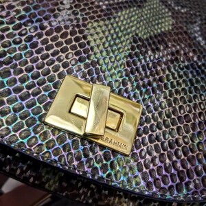 May include: A handbag featuring a textured, iridescent snakeskin pattern in shades of green, black, and purple. The bag is secured with a rectangular, polished gold-tone metal turn-lock closure that is clearly engraved with the brand name BRAHMIN.