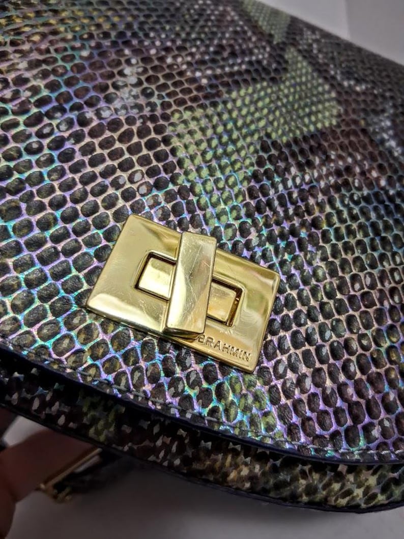 May include: A handbag featuring a textured, iridescent snakeskin pattern in shades of green, black, and purple. The bag is secured with a rectangular, polished gold-tone metal turn-lock closure that is clearly engraved with the brand name BRAHMIN.