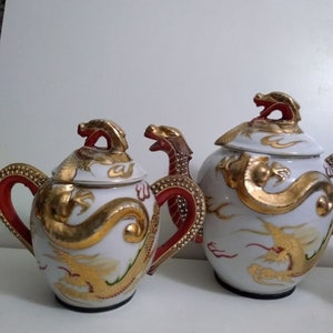 May include: Two white ceramic teapots with gold dragon designs and red accents. The teapots have gold dragon-shaped handles and lids. The dragon design is painted in gold. The teapots are on a white surface.