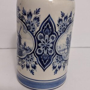 May include: A cylindrical ceramic mug with a blue and white floral and landscape design. The mug features a central floral motif and framed scenes of windmills and buildings. The rim and base are accented with blue bands.
