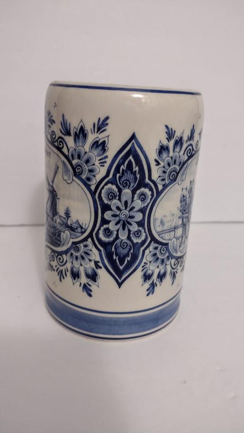 May include: A cylindrical ceramic mug with a blue and white floral and landscape design. The mug features a central floral motif and framed scenes of windmills and buildings. The rim and base are accented with blue bands.
