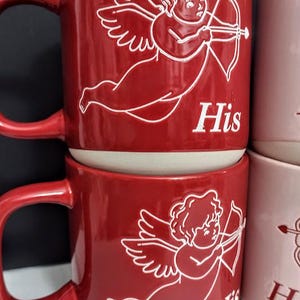 May include: Red ceramic mugs with white cherub and bow and arrow illustrations. The word "His" is printed on the front of the mug. The mugs have a curved handle and a white interior. The mugs are stacked on top of each other.