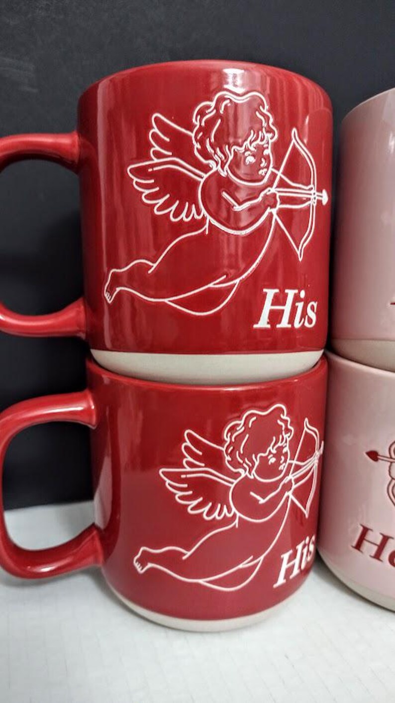 May include: Red ceramic mugs with white cherub and bow and arrow illustrations. The word "His" is printed on the front of the mug. The mugs have a curved handle and a white interior. The mugs are stacked on top of each other.
