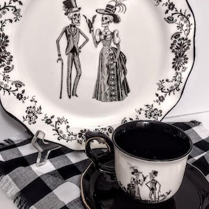 May include: A white ceramic plate with a scalloped edge and black floral border, featuring a central illustration of two skeletons in formal attire. A matching teacup and saucer set with a black interior and a skeleton couple graphic.