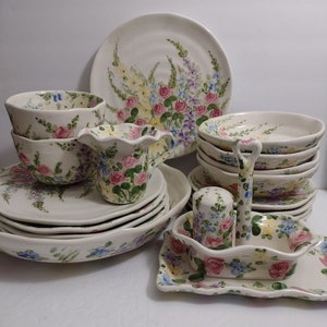 May include: A collection of white ceramic dinnerware with a hand-painted floral design. The set includes plates, bowls, a creamer, a salt and pepper shaker, and a serving dish. The floral pattern features pink, blue, and yellow flowers with green leaves.