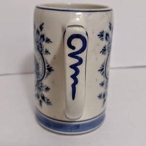 May include: A white ceramic mug with blue floral patterns and a blue handle design. The mug has a blue band at the base and rim. The handle features a blue, stylized swirl design. The mug is a classic design.