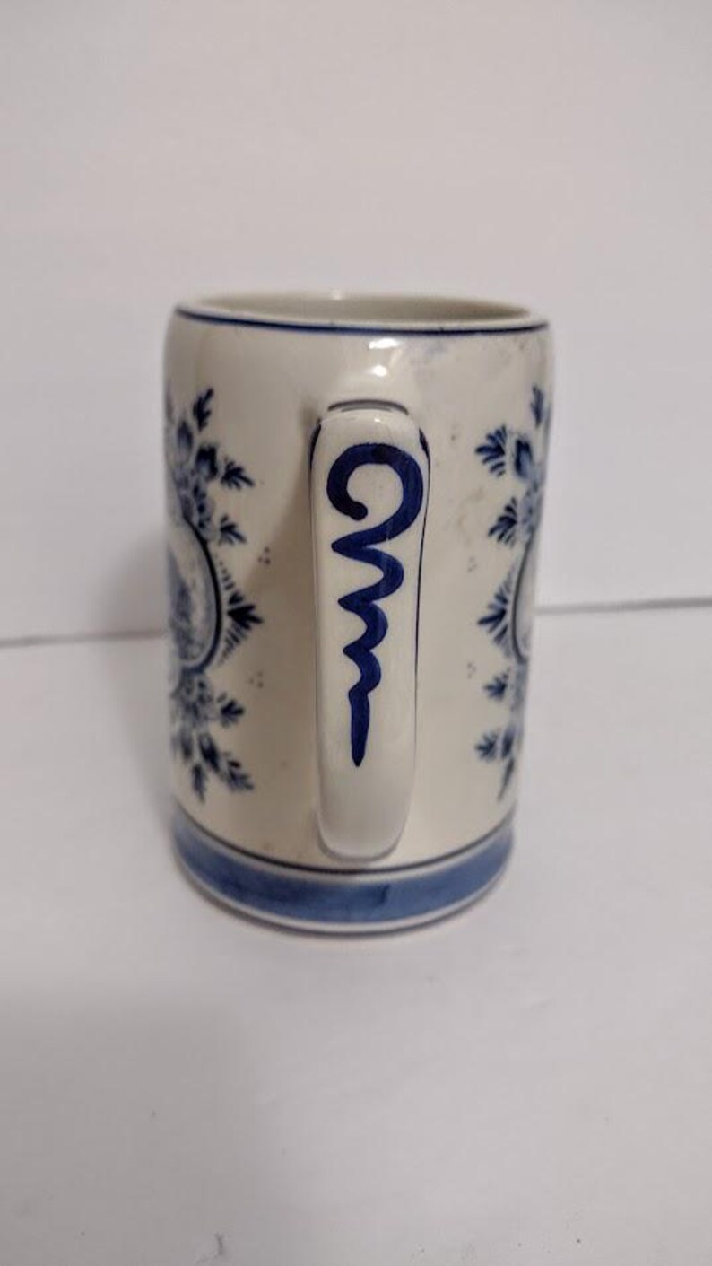 May include: A white ceramic mug with blue floral patterns and a blue handle design. The mug has a blue band at the base and rim. The handle features a blue, stylized swirl design. The mug is a classic design.