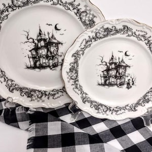 May include: Two white plates with black haunted house illustrations and ornate black borders. The plates have scalloped edges with gold trim. A black and white checkered cloth is beneath the plates.