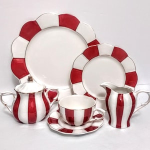 May include: A ceramic dinnerware set featuring a red and white striped pattern. The collection includes a large dinner plate, a smaller salad plate, a lidded sugar bowl with two handles, a teacup on a matching saucer, and a creamer pitcher.