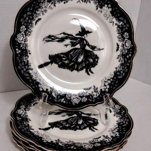 May include: A stack of white ceramic plates with scalloped edges and a gold-trimmed rim. The border features a black floral pattern. The center of each plate displays a black illustration of a figure wearing a pointed hat and riding a broomstick.