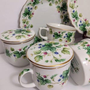May include: White ceramic teacups, saucers, and plates with a gold trim. The set features a green leaf and berry design. The teacups have lids. The plates have a scalloped edge.