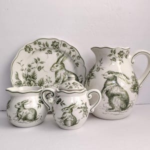 May include: A white ceramic tea set with a green rabbit and floral design. The set includes a plate, a pitcher, a sugar bowl, and a creamer. The plate has a scalloped edge.