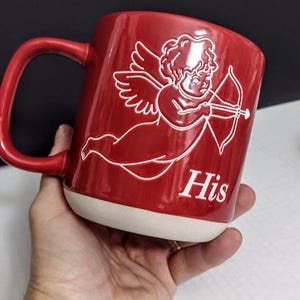 May include: Red ceramic mug with a white base and handle. The mug features a white illustration of Cupid with a bow and arrow, and the word "His" in white lettering.