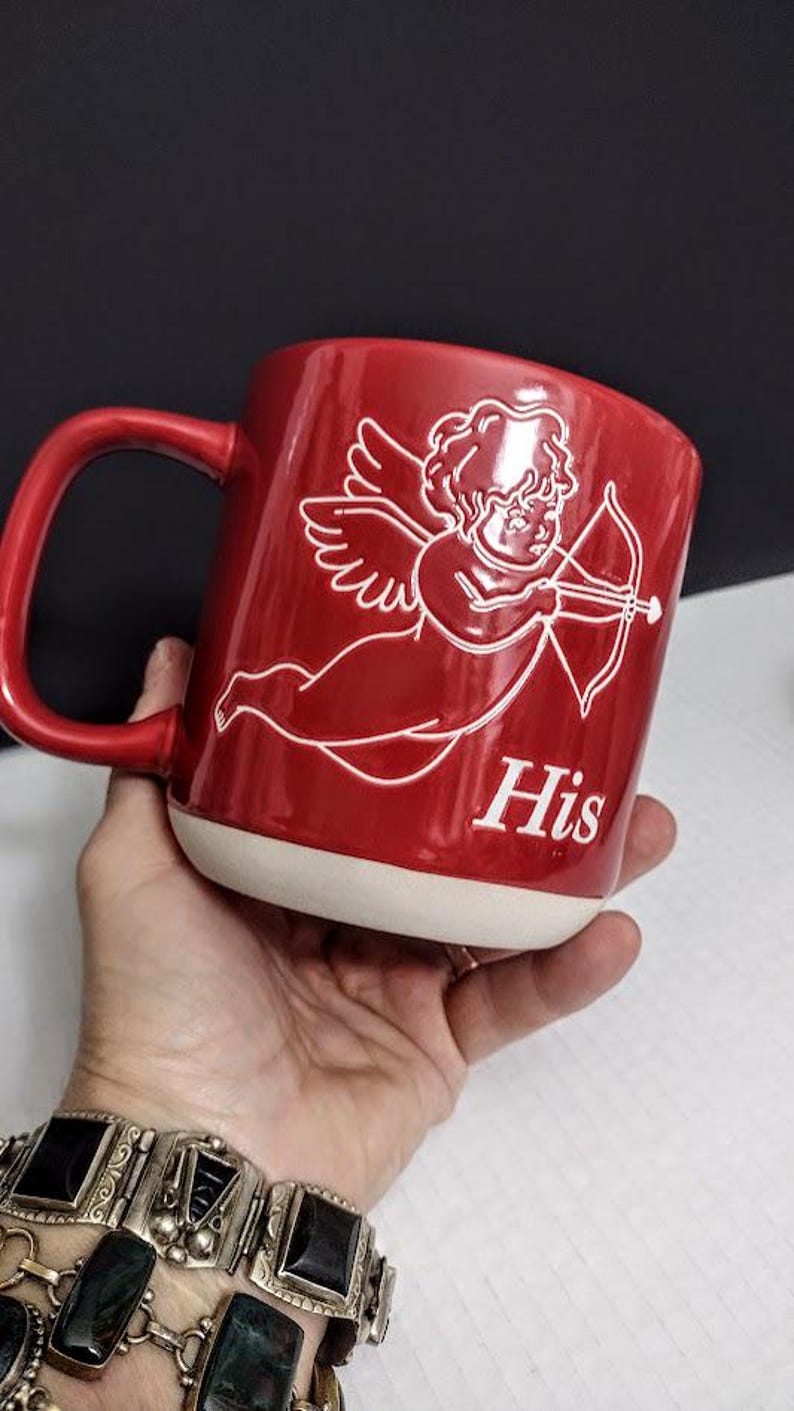 May include: Red ceramic mug with a white base and handle. The mug features a white illustration of Cupid with a bow and arrow, and the word "His" in white lettering.
