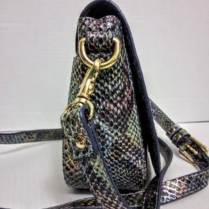 May include: A crossbody bag featuring a multi-coloured snakeskin pattern in shades of green, blue, and purple. The bag includes gold-tone metal hardware, a detachable strap with a gold-tone lobster clasp, and an adjustable buckle on the strap.
