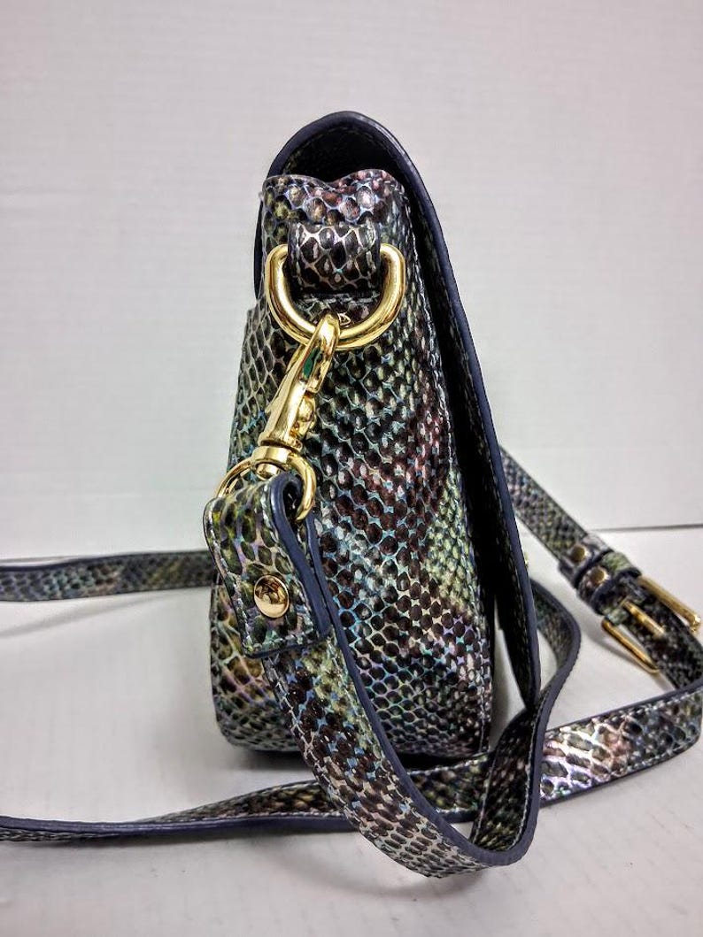 May include: A crossbody bag featuring a multi-coloured snakeskin pattern in shades of green, blue, and purple. The bag includes gold-tone metal hardware, a detachable strap with a gold-tone lobster clasp, and an adjustable buckle on the strap.