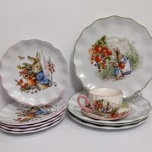 May include: A set of white ceramic plates and a teacup with a floral and bunny design. The plates have scalloped edges with pink or green trim. The teacup has a pink handle and a matching saucer.