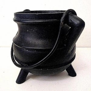 May include: A matte black cast iron cauldron featuring a rounded body, a wide rim, and three short, sturdy legs. The vessel includes a thick, curved metal handle attached to two side lugs, allowing it to be carried or suspended upright.