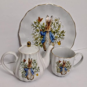 May include: A white ceramic tea set featuring a Peter Rabbit design. The set includes a teapot, a teacup, and a plate, all adorned with illustrations of Peter Rabbit, butterflies, and floral accents. The edges are trimmed with gold.