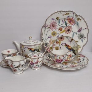 May include: A complete floral tea set with a teapot, cups, saucers, creamer, sugar bowl, and serving plates. The set features a bird and floral design in pink, yellow, and green on a white background. The plates have a scalloped edge.
