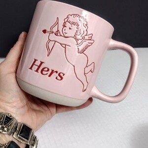 May include: A pink ceramic mug with a white base and handle. The mug features a red illustration of Cupid with a bow and arrow, and the word "Hers" in red lettering. The mug is held in a hand.
