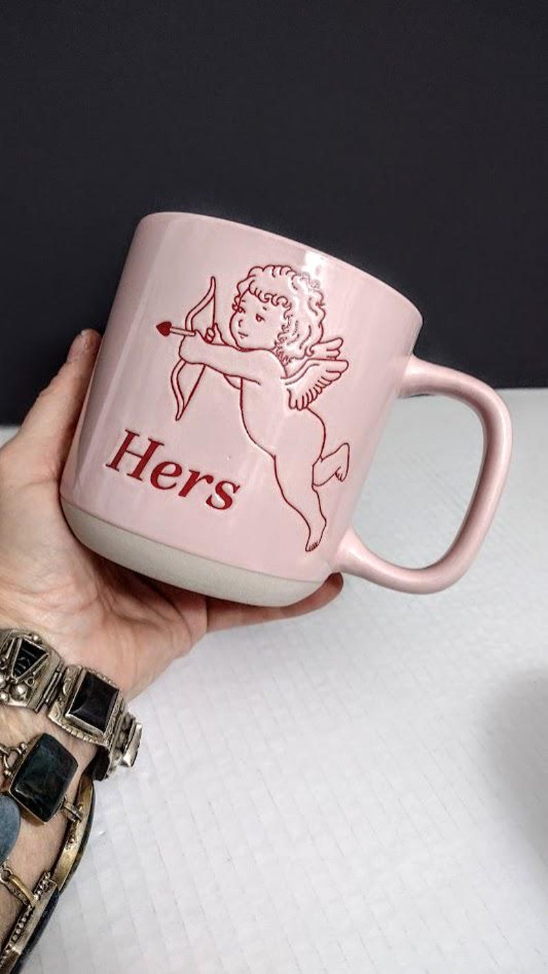 May include: A pink ceramic mug with a white base and handle. The mug features a red illustration of Cupid with a bow and arrow, and the word "Hers" in red lettering. The mug is held in a hand.