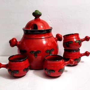 May include: A bright red ceramic soup tureen with a matching lid and four small handled bowls. Each piece features a dark green three-leaf clover pattern and a dark brown rim. The tureen has two side handles and a green knob on the lid.