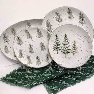 May include: A set of white ceramic plates with a speckled design and green Christmas tree illustrations. The plates are displayed on clear stands, with a green fabric napkin featuring "Merry Christmas" text.