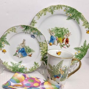 May include: A white ceramic tea set featuring illustrations of rabbits in blue and red jackets, surrounded by ferns and flowers. The set includes a mug with a gold handle and rim, and two plates with scalloped edges. A colorful Easter-themed napkin is in the foreground.