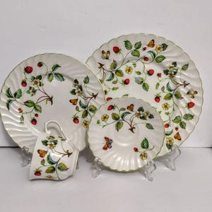 May include: A white ceramic dinnerware set featuring a scalloped edge and a repeating pattern of red strawberries, green leaves, yellow flowers, and small butterflies. The set includes a large dinner plate, a smaller plate, and a teacup.