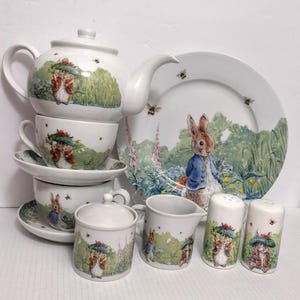 May include: A white ceramic tea set featuring illustrations of Peter Rabbit and other characters. The set includes a teapot, cups, saucers, a sugar bowl, creamer, a plate, and salt and pepper shakers. The illustrations feature green, brown, and blue colors.
