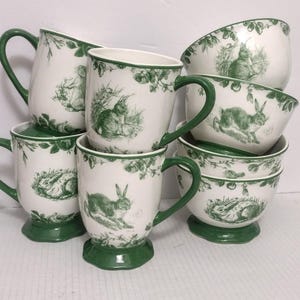 May include: A collection of white ceramic mugs and bowls featuring a green transferware pattern of rabbits and foliage. Each piece has a green base and handle, with detailed illustrations of rabbits in various poses amidst leafy branches.