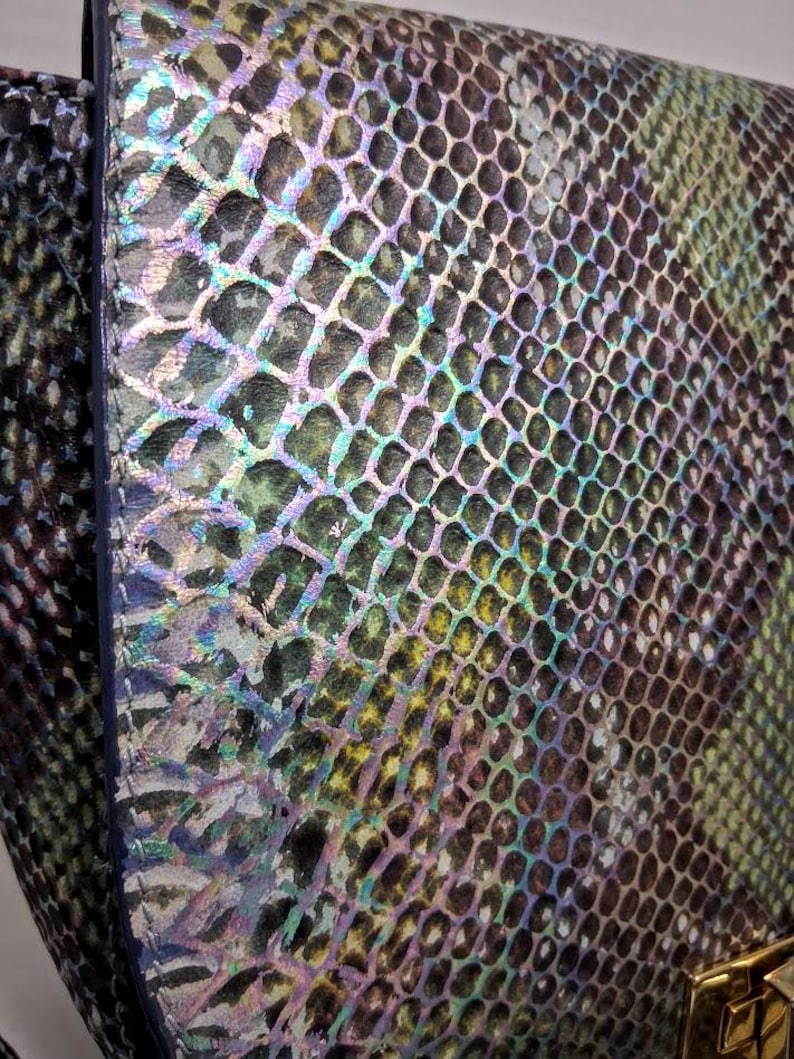 May include: A handbag featuring a textured snakeskin pattern with an iridescent finish. The surface displays a gradient of green, black, and purple hues with a metallic sheen. A gold-toned metal clasp is visible at the bottom right corner.