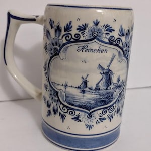 May include: A white ceramic beer stein with blue floral and windmill designs. The stein features a handle and a blue band at the base. The word "Heineken" is displayed on the front.