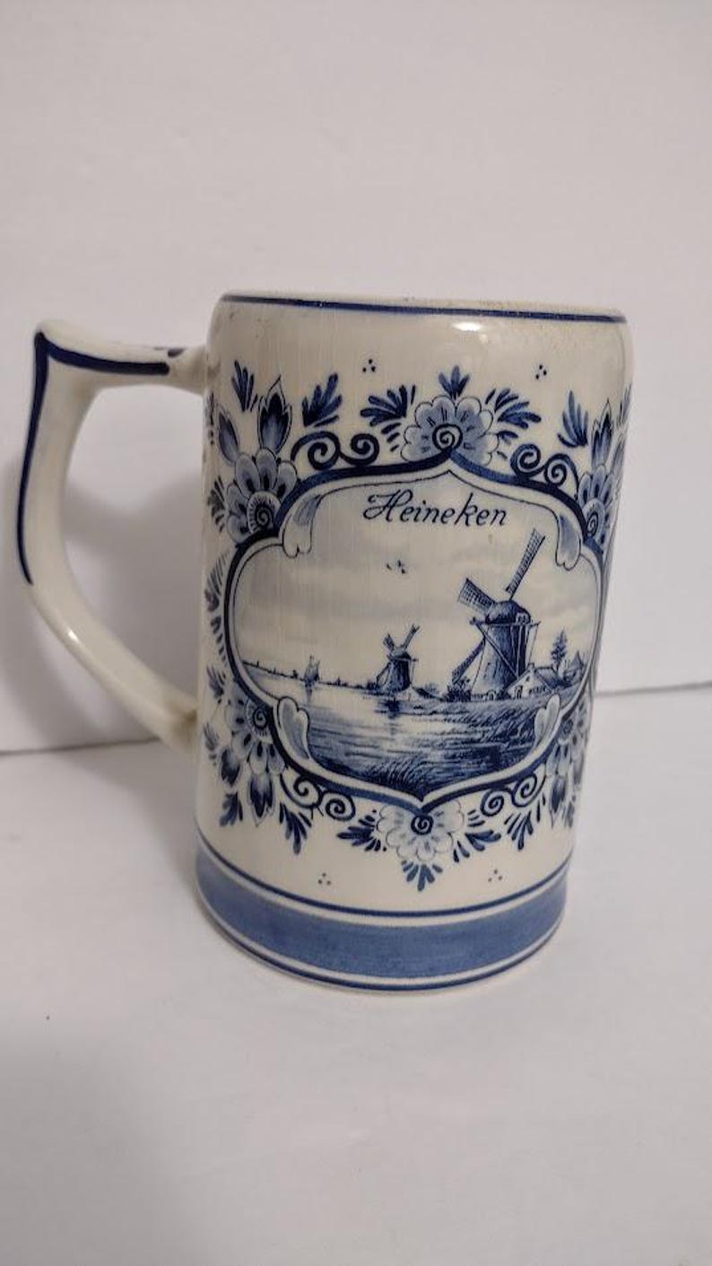 May include: A white ceramic beer stein with blue floral and windmill designs. The stein features a handle and a blue band at the base. The word "Heineken" is displayed on the front.