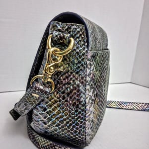May include: A crossbody bag featuring a snakeskin pattern with iridescent hues of blue, green, and purple. The bag includes a gold-toned metal lobster clasp attachment connecting the adjustable strap to the side of the structured, rounded body.