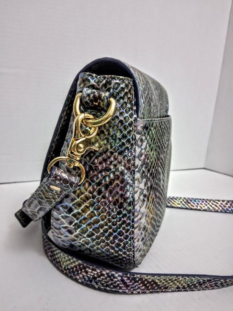 May include: A crossbody bag featuring a snakeskin pattern with iridescent hues of blue, green, and purple. The bag includes a gold-toned metal lobster clasp attachment connecting the adjustable strap to the side of the structured, rounded body.
