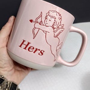 May include: A pink ceramic mug with a white base and a curved handle. The mug features a red illustration of Cupid with a bow and arrow, and the word "Hers" in red. The mug is held by a person.
