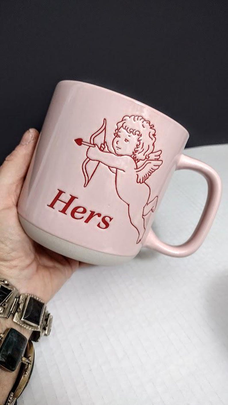 May include: A pink ceramic mug with a white base and a curved handle. The mug features a red illustration of Cupid with a bow and arrow, and the word "Hers" in red. The mug is held by a person.