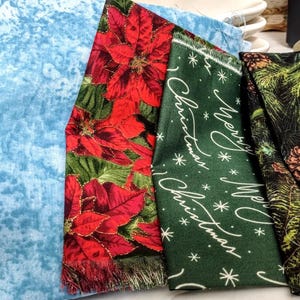 May include: A collection of folded fabrics with various patterns. Includes a red and black plaid, a light blue textured fabric, a red poinsettia print, a green fabric with "Merry Christmas" text, and a black fabric with pine cones.