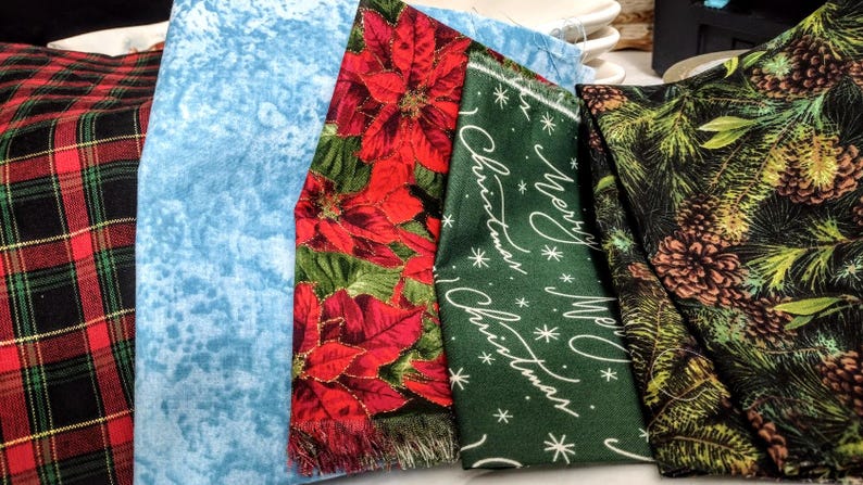 May include: A collection of folded fabrics with various patterns. Includes a red and black plaid, a light blue textured fabric, a red poinsettia print, a green fabric with "Merry Christmas" text, and a black fabric with pine cones.