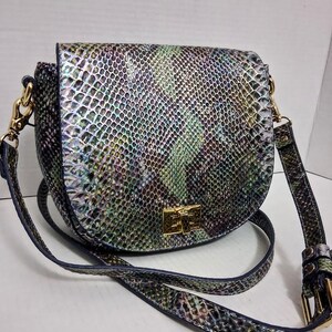May include: A saddle-shaped crossbody bag featuring an iridescent snakeskin pattern in shades of green, purple, and silver. The bag includes a gold-tone rectangular turn-lock closure on the front flap and an adjustable shoulder strap with gold hardware.