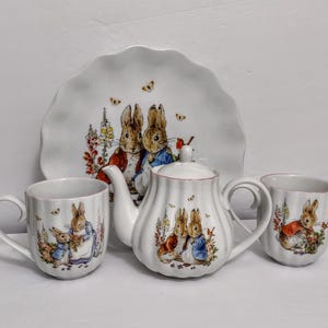 May include: A white ceramic tea set featuring illustrations of rabbits in a garden setting. The set includes a teapot, two mugs, and a plate with a scalloped edge. The teapot and mugs have a pink trim. The plate measures approximately 10 inches in diameter.