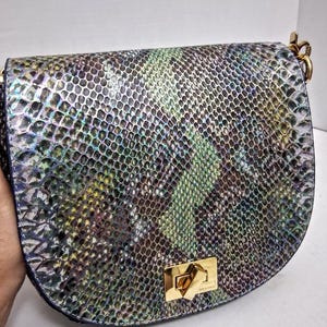 May include: A saddle-shaped shoulder bag featuring a textured snakeskin pattern in iridescent shades of green, blue, purple, and silver. The bag includes a gold-toned metal turn-lock closure on the front flap and gold-toned hardware attachments.